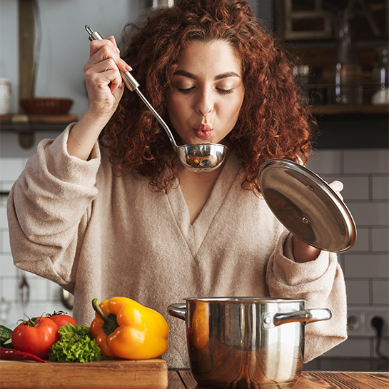 A woman in the kitchen holding a pot lid and a spoon, ready to taste the soup