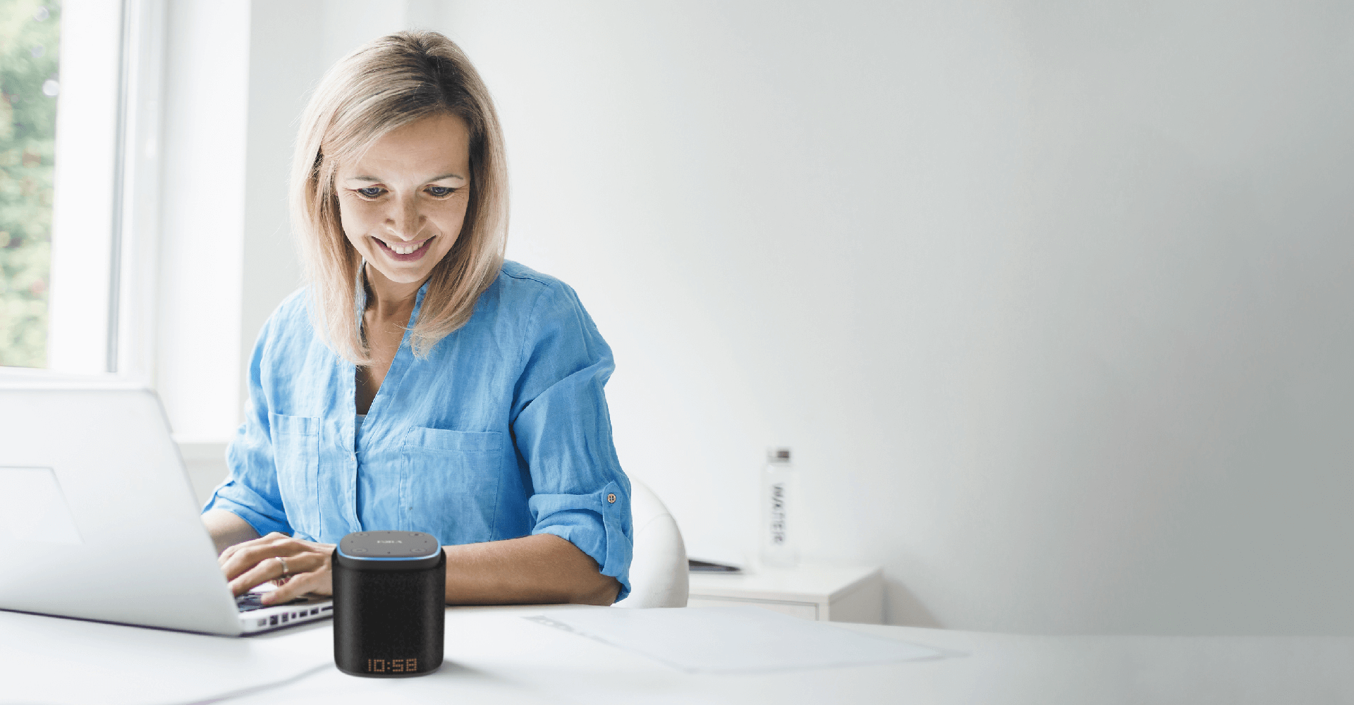 A woman at office desk using computer, talking to iFORA IntelliSpeaker