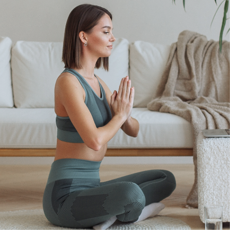 A woman sitting on the floor at home, hands folded in meditation