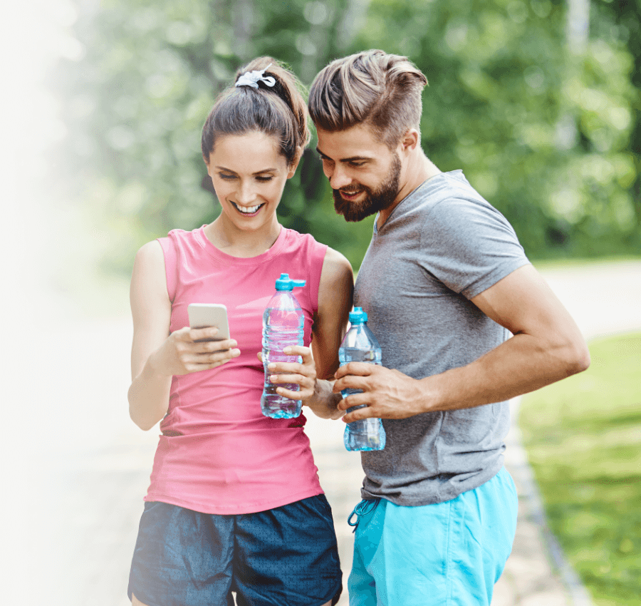 A smiling couple holding water bottles and a phone outdoors, ready for sports