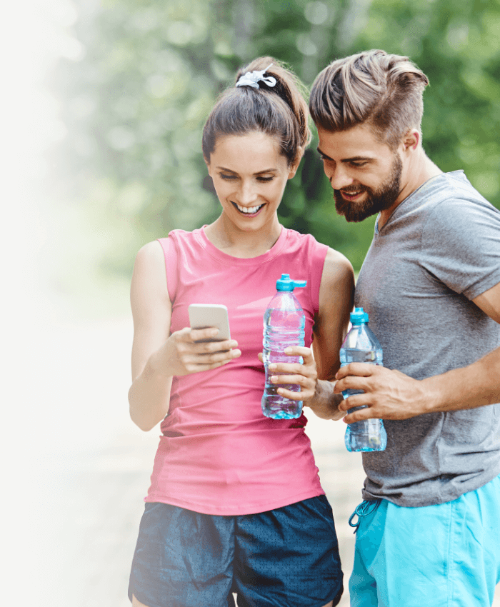 A smiling couple holding water bottles and a phone outdoors, ready for sports